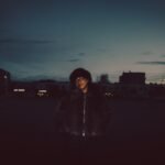Model in black jacket standing outdoors at dusk